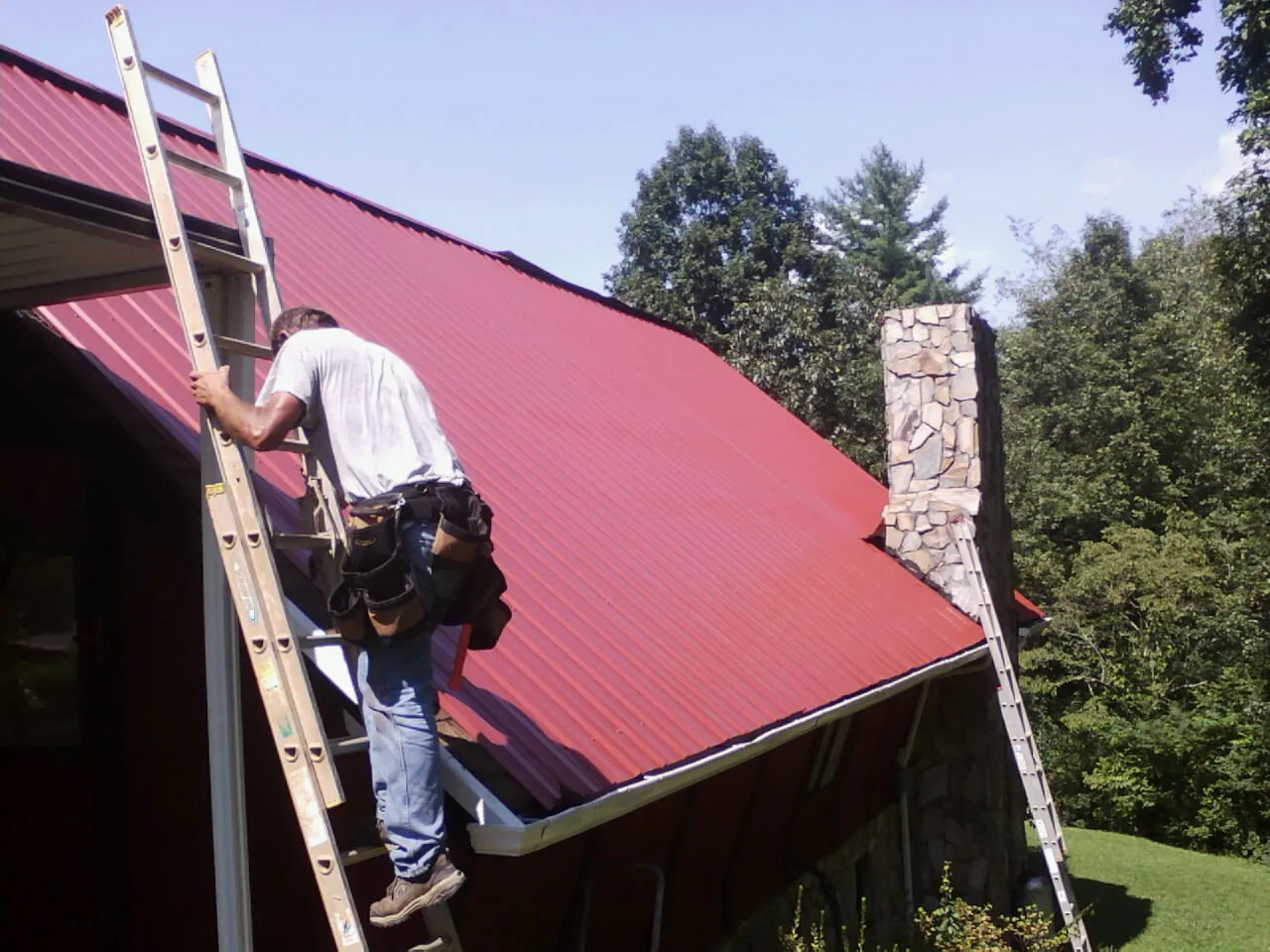 Roofer on a ladder beside a red metal roof and stone chimney — Soffit & Fascia Repair by Roofing Brothers in Cokato